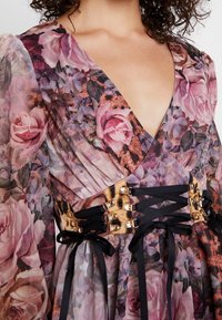 Robe fleurie en tissu transparent et texturé avec un décolleté en V profond et une taille cintrée ornée de matériaux dorés et de rubans noirs.