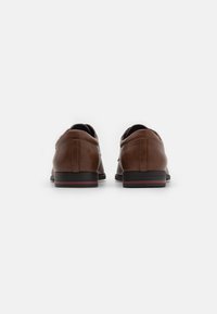 Chaussures en cuir marron avec une texture lisse, une forme arrondie et un talon bas. Présentent des accents rayés noirs et rouges sur la semelle.