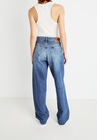 Osoba v bílém bezrukávovém topu a volných modrých džínách značky Lee, zobrazena zezadu na jednobarevném bílém pozadí.