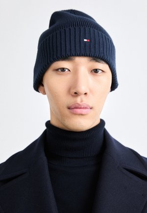 Marineblaue gerippte Strickmütze mit einem kleinen weißen, roten und blauen Logodetail. Das Modell trägt einen passenden marineblauen Rollkragenpullover und einen Mantel.