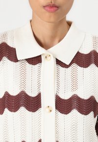 Cardigan lavorato a maglia con un motivo ondulato crema e rosso scuro, dotato di colletto a polo e chiusura centrale con bottone. Tessuto leggero e testurizzato.