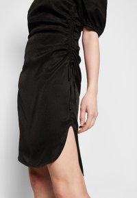 Czarna sukienka marszczona z krótkim rękawem typu bufka i bocznym rozcięciem, widziana od połowy uda do tuż poniżej ramienia na stojącej osobie.