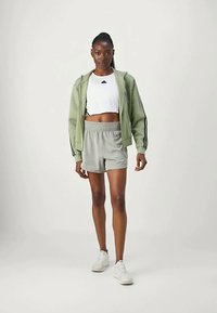 Γυναίκα που προχωράει φορώντας ανοιχτό πράσινο φούτερ με φερμουάρ, λευκό κοντομάνικο μπλουζάκι, γκρι σορτσάκι και λευκά αθλητικά παπούτσια μπροστά σε απλό φόντο.