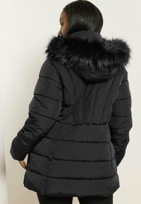 Svart puffjacka med huva i fuskpäls, med en kviltad design och långa ärmar, som sträcker sig till mid-lår med en figurnära silhuett.