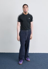 Zwarte Adidas T-shirt met witte strepen, losse marineblauwe gestreepte broek en paarse suède sneakers met gumzolen. Staande tegen een neutrale achtergrond.