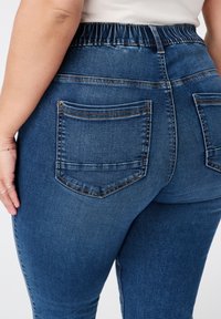 Jean en denim bleu foncé avec une taille élastique, dotés de deux poches arrière à patch et de détails de couture contrastants. Texture lisse.