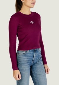 Dlouhý rukáv, cropped top v tmavé vínové barvě s žebrovanou texturou. Na hrudi je bílý logo Calvin Klein. Kombinováno s modrými džínami.