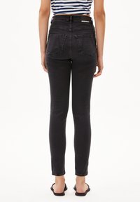 Jeans skinny de tiro alto en negro con una textura suave, que presentan dos bolsillos traseros y una etiqueta de logo en la cinturilla. Combinados con sandalias.