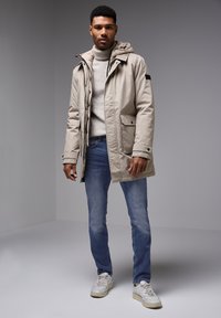 Beige voděodolná parka s kapucí, zapínáním na zip a bočními kapsami, přes světlou šedou rolák, spárovaná s modrými dž ínami a bílými teniskami.