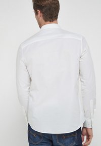 Chemise blanche à manches longues avec col classique, poignets à boutons et couture de yoke arrière. Tissu en coton lisse avec une coupe régulière, associée à un jean bleu foncé.