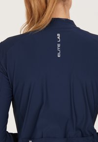 Giacca sportiva navy con colletto alto, texture liscia e logo "ELITE LAB" in bianco sul retro, caratterizzata da un design slim fit.