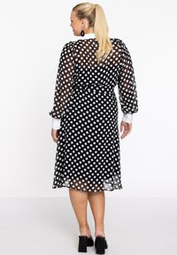 Schwarz-weißes Pünktchenkleid mit durchsichtigen langen Ärmeln, tailliertem Bund und weißem Kragen. Ausgestellter Saum des Rocks, kombiniert mit Schuhen mit Blockabsatz.