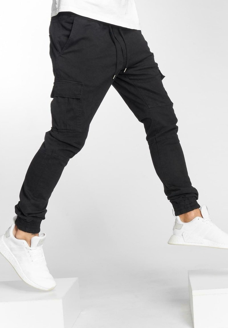 Personne portant un pantalon cargo jogger noir et des baskets blanches marchant entre deux plateformes blanches sur un fond uni.