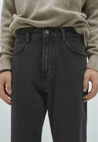 Czarne dżinsy o prostym kroju z pięcioma kieszeniami, z zapinaniem na guzik i subtelnymi detalami w przeszyciach. Noszone z beżowym swetrem w prążki.