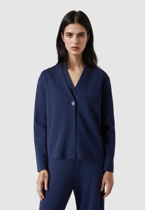 Junge Frau mit glattem dunklem Haar, die eine marineblaue V-Ausschnitt-Strickjacke mit Knöpfen und passende Hose trägt, steht vor einem einfarbigen Hintergrund.