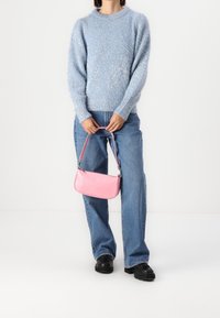 Personne portant un pull en laine léger bleu clair et un jean bleu, tenant un petit sac à main rose avec des chaussures noires sur un fond blanc.