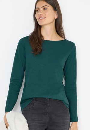 Top de manga larga en color verde oscuro, hecho de una tela suave, con un escote barco y un dobladillo ligeramente curvado. Combinado con unos jeans gris oscuro.