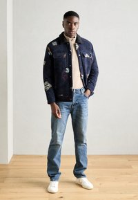 Ονειρεμένο navy σακάκι με κεντημένα λουλουδάτα σχέδια, μπεζ φερμουάρ πουκαμίσα, μπλε τζιν και λευκά αθλητικά παπούτσια, σε ανοιχτό φόντο.