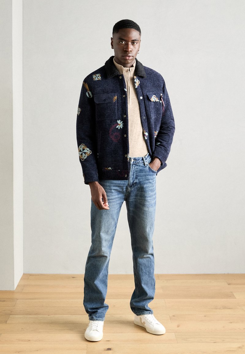 Ονειρεμένο navy σακάκι με κεντημένα λουλουδάτα σχέδια, μπεζ φερμουάρ πουκαμίσα, μπλε τζιν και λευκά αθλητικά παπούτσια, σε ανοιχτό φόντο.