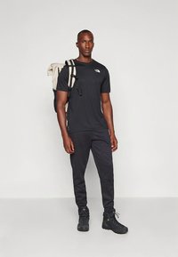 Black short-sleeve t-shirt with logo, black tapered pants, and dark boots. Model carries a beige backpack with dark straps. Smooth texture.