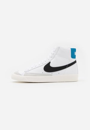 Biały, wysok-topowy sneaker Nike z czarnym logo, niebieską zakładką na pięcie i kremową podeszwą, pokazany z boku na białym tle.