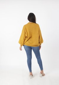 Maglione lavorato a maglia oversize color senape con maniche lunghe a palloncino; polsini e orlo a costine; abbinato a jeans aderenti blu e sandali trasparenti con tacco.