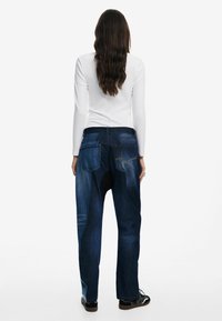 Denimjeans mit mittlerer Waschung und ausgewaschenen Stellen, mit lockerem Schnitt, fünf Taschen und Kontrastnähten. Getragen mit einem weißen Langarm-Shirt.