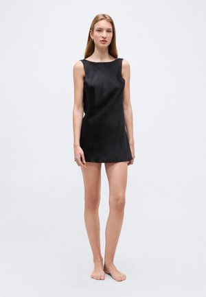 Fiatal nő mezítláb áll, ujjatlan fekete miniruha van rajta, egy egyszerű világosszürke háttér előtt.