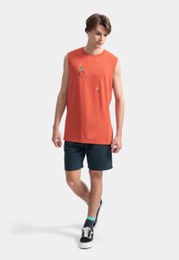 Chemise sans manches orange avec des designs graphiques, associée à un short bleu foncé ; le modèle porte des baskets noires et des chaussettes turquoise.