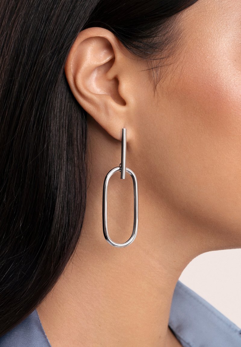 Gros plan sur l’oreille d’une femme portant une boucle d’oreille pendante ovale allongée en argent avec une barre, cheveux raides foncés et peau brun clair.