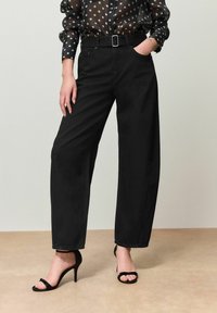 Schwarze, weit geschnittene Baumwollhose mit hohem Bund, ausgestattet mit Gürtel und Seitentaschen, kombiniert mit schwarzen, Riemchen-Highheels.