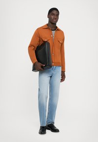 Brun jakke med to brystlommer, grå t-shirt, lyseblå jeans og sorte sko. Holder en stor sort lædertaske. Ensfarvet baggrund.