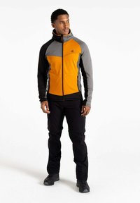Orange- und graue Jacke mit Reißverschluss und Kapuze, mit einem geometrischen Design. Schwarze Hose und robuste Schuhe runden das Outfit ab.