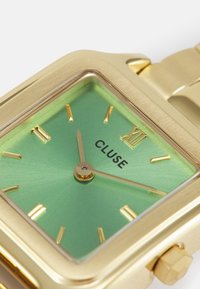 Montre rectangulaire dorée avec un cadran dégradé vert. Présente des index et aiguilles dorés, avec une couronne sur le côté et le logo "CLUSE".