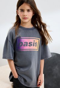 Niña con cabello largo y castaño, vistiendo una camiseta gris holgada con un logo degradado de "oasis" en púrpura y naranja, de pie en interiores con las manos en los bolsillos.