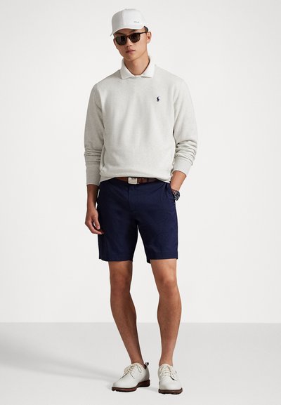 Jonge man met een witte pet, zonnebril, lichtgrijze trui over een witte overhemd, marineblauwe shorts, witte schoenen en een horloge, staande tegen een witte achtergrond.