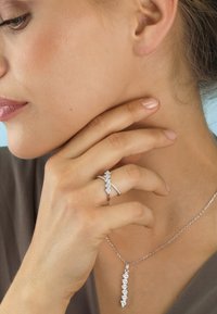 Woman wearing a silver ring with diamonds and a matching diamond pendant necklace, touching her neck gently.