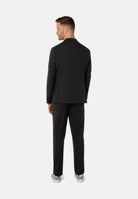 Veste de costume noire à coupe slim, avec un col à revers cranté et une fente unique, associée à un pantalon assorti et des baskets gris clair.