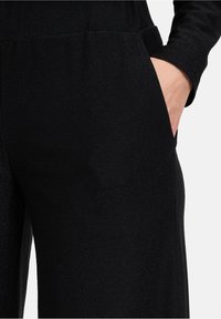 Zwarte jumpsuit van gestructureerde stof, met een relaxte pasvorm, zijzakken en subtiele glans. Lange mouwen en elastische tailleband.