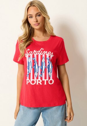 Blondine die een rood T-shirt draagt met blauwe sardine illustraties en de woorden "Sardines PORTO", gecombineerd met lichtblauwe spijkerbroek.