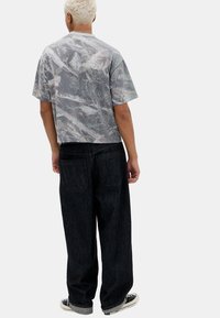 Afskåret grå t-shirt med camouflage-mønster på en struktureret stof, parret med løse, mørke denimjeans og casual sneakers.