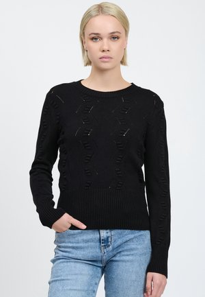Donna bionda con capelli corti che indossa un maglione nero a maglia testurizzata e jeans azzurri chiari, con una mano in tasca, su uno sfondo bianco uniforme.