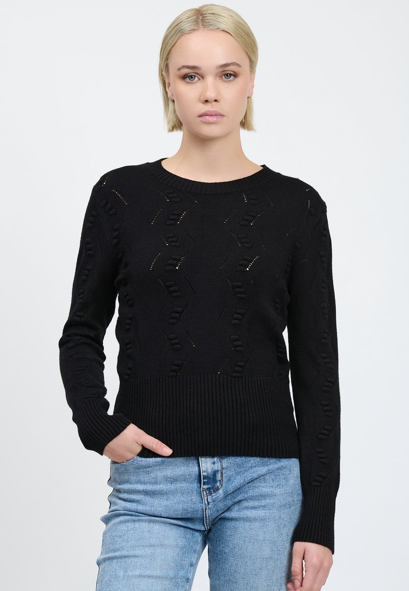 Donna bionda con capelli corti che indossa un maglione nero a maglia testurizzata e jeans azzurri chiari, con una mano in tasca, su uno sfondo bianco uniforme.