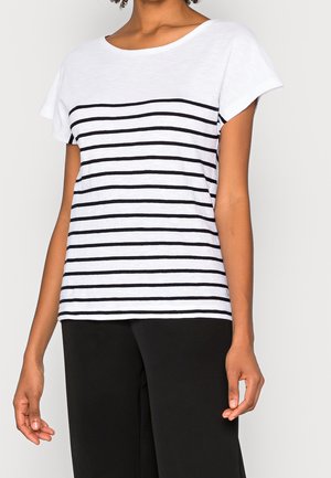 Person trägt ein weißes kurzärmeliges Oberteil mit schwarzen horizontalen Streifen und schwarze Hose, steht vor einem schlichten Hintergrund.
