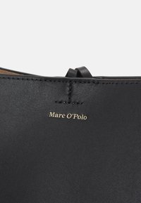 Szczegół czarnego skórzanego buta, z przeszytymi krawędziami i złotym logo "Marc O'Polo" na gładkiej fakturze. Zawiera małą skórzaną pętlę.