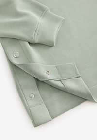 Manche et ourlet de vêtement vert clair avec boutons-pression, montrant un côté partiellement ouvert pour une fermeture et un ajustement faciles.