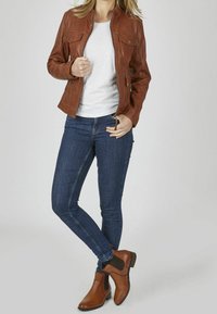 Giacca di pelle marrone con zip frontale, due tasche sul petto e design aderente. Abbinata a jeans skinny blu scuro e stivali alla caviglia marroni.