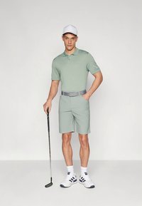 Μπλούζα πόλο με κάθετες ρίγες σε ανοιχτό πράσινο, συνδυασμένη με matching σορτς, γκρι ζώνη, λευκό καπέλο και λευκά παπούτσια με λεπτομέρειες. Κλαμπ γκολφ στο χέρι.