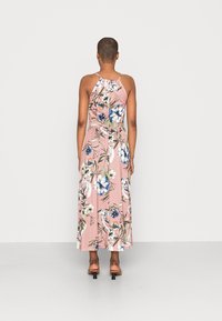 Femme debout, vêtue d'une robe maxi rose sans manches avec un motif floral bleu, blanc et vert, vue de dos, sur un fond uni.