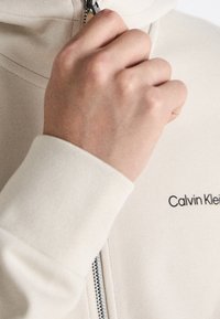 Felpa beige con zip, dal tessuto morbido, caratterizzata da una zip argento e dal logo nero "Calvin Klein". Mano visibile che afferra la zip.